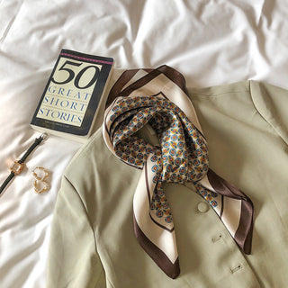 Elegant silk scarf with geometric pattern in red, blue and brown border, styled on beige blazer with book and accessories on white bedding