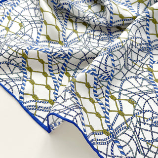 Silk scarf with white background featuring blue rope-like geometric pattern and olive green accents, showing luxurious fabric texture and blue trim