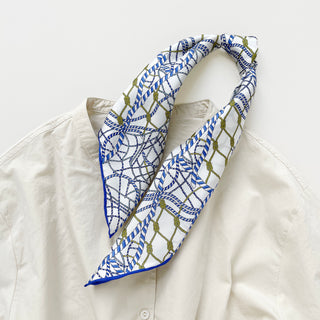 White silk bandana with blue nautical rope pattern and olive knot details, styled around collar of cream shirt, showcasing hand-rolled blue edge
