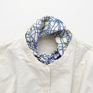 White and blue geometric silk bandana with nautical rope pattern and gold accents, styled around the collar of a cream button-up shirt