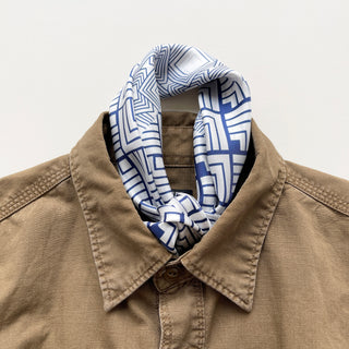 White silk bandana with blue geometric patterns tied stylishly around the collar of a tan casual shirt, showcasing versatile styling