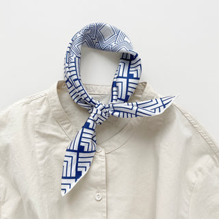 White silk bandana with blue geometric pattern tied stylishly around collar of cream button-up shirt, showcasing modern unisex design