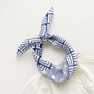 White silk bandana with blue geometric pattern tied in a knot, displayed on light background showing hand-rolled edges and modern design