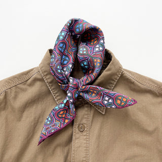 Plum purple silk neckerchief with colorful geometric pattern in blue and orange, tied around collar of tan shirt, showcasing hand-rolled edges