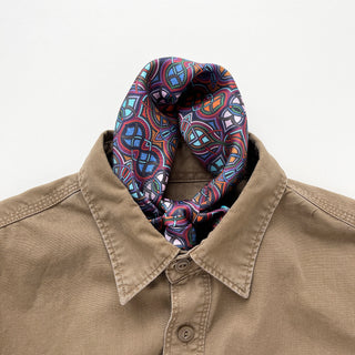 Plum purple silk neckerchief with geometric pattern in blue and orange, styled casually with a tan button-up shirt, showing luxurious texture and vibrant design