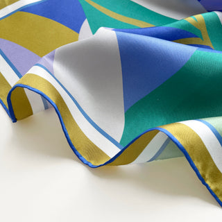 Luxurious silk scarf featuring abstract geometric pattern in blue, teal, mustard yellow, and lilac with elegant hand-rolled blue edges