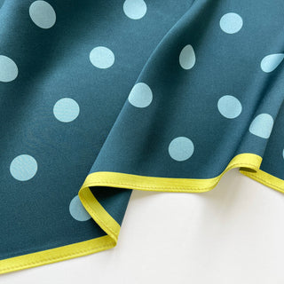 Teal green silk square scarf with light blue polka dots and bright mustard yellow border, elegantly folded showing luxurious sheen and texture