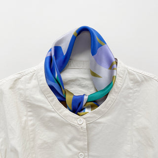 Abstract lotus silk scarf in vibrant blue, green, lilac, and mustard yellow, styled as a neckerchief with a white button-up shirt