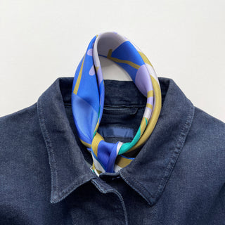 Blue abstract lotus silk scarf with vibrant mustard yellow, lilac and teal accents, styled as a neckerchief on dark denim shirt collar