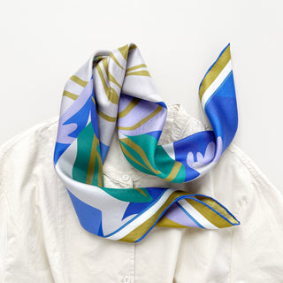 Luxurious abstract lotus silk scarf in vibrant blue, teal, lilac, and mustard yellow patterns, elegantly draped around a white shirt collar.