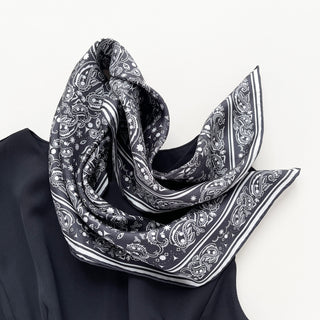 Black silk scarf with white paisley pattern and striped border, elegantly draped to showcase its luxurious texture and hand-rolled edges
