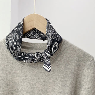Black and white paisley silk scarf tied elegantly around neckline of light gray sweater, showcasing intricate pattern and hand-rolled edges