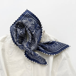 Navy silk scarf with white paisley pattern and striped hand-rolled edges, styled as a neckerchief on a cream button-up shirt