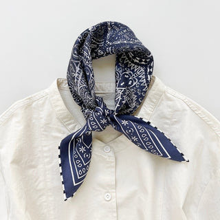Navy silk bandana with white paisley pattern tied around collar of cream shirt, featuring hand-rolled edges and decorative striped border