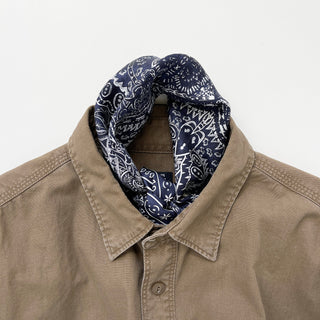 Navy silk bandana with white paisley pattern styled as a neckerchief with a tan button-up shirt, displaying the scarf's intricate design.