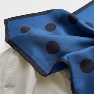 Yale blue silk scarf with black polka dots and hand-rolled navy border, displaying luxurious sheen and elegant drape on light surface.