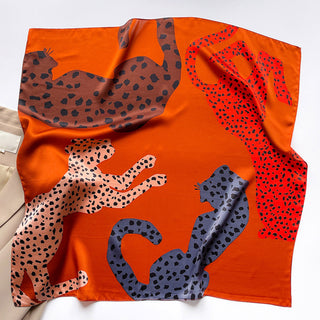 Burnt orange silk scarf with colorful leopard silhouettes in brown, pink, red, and blue, featuring spotted pattern designs on a square 65cm bandana