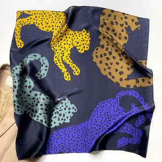 Square silk scarf with navy blue background featuring colorful leopard silhouettes in yellow, bronze, teal, and indigo, showcasing luxurious sheen