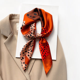 Square silk scarf with burnt orange, brown and pink leopard print pattern, styled in a knot on beige blazer with white card background