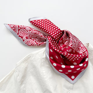 Red silk bandana scarf with combined paisley and polka dot patterns, featuring white designs on vibrant red fabric with silver trim, tied stylishly on a white shirt.
