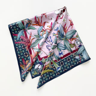Square silk scarf with pink botanical print featuring blue-green leaves, colorful flowers, and navy border with geometric pattern. Luxurious sheen on folded display.