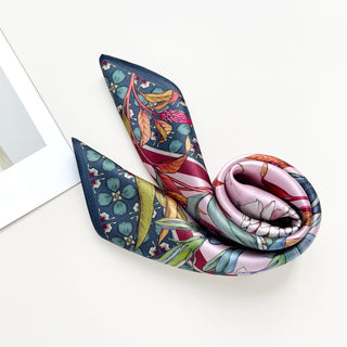 Silk bandana scarf with botanical floral print featuring pink, dark green, and gold tones, elegantly displayed with gentle folds showing its luxurious texture