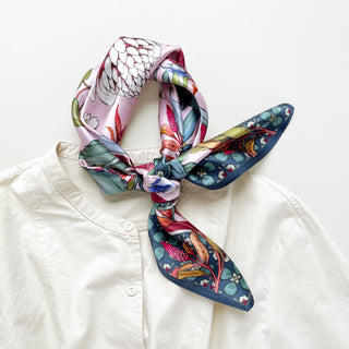 Pink silk bandana scarf with botanical floral print tied at neck of white blouse, featuring dark green border and vibrant flower motifs
