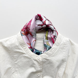 Pink silk bandana scarf with botanical floral print in vibrant pinks, whites, and dark green, styled around the collar of a cream blouse
