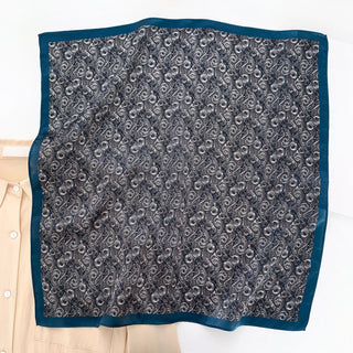 Square silk neckerchief with deep blue border and navy-white wave pattern interior, displayed flat beside light beige garment