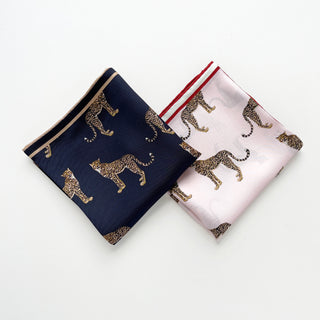 Two luxury silk scarves with leopard print pattern - one in navy blue with cream border and one in pink with red trim, displayed side by side