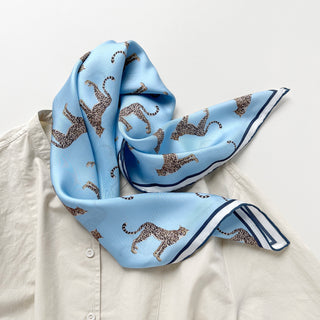 Sky blue silk scarf with leopard print pattern and navy border, elegantly draped over a light beige shirt, showcasing its luxurious twill texture