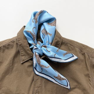 Sky blue silk neckerchief with leopard print pattern tied around collar of tan jacket, featuring luxurious sheen and navy border trim