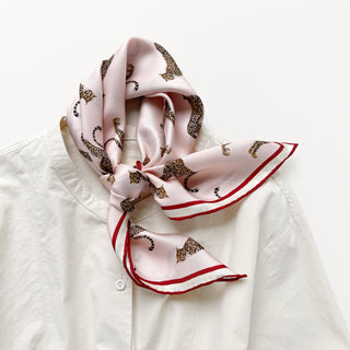 Pink silk scarf with leopard print pattern and red border, elegantly tied around collar of white shirt, showcasing luxurious hand-rolled edges
