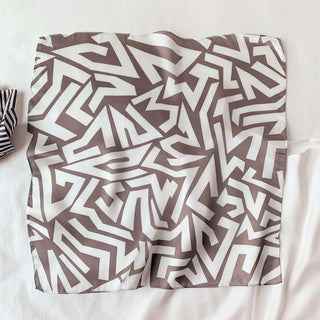 Square silk bandana scarf with geometric zigzag pattern in taupe brown on creamy white background, displaying luxurious sheen