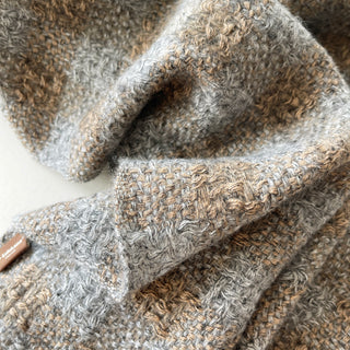 A close-up of a soft, textured knit cashmere scarf  in a heather mix of grey, beige, and brown tones, draped to show its tactile weave and colour variation. 