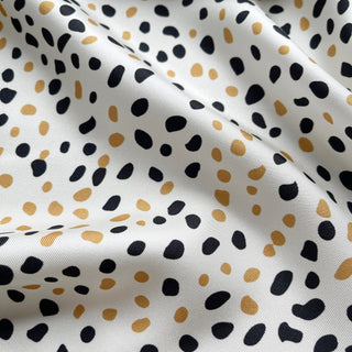 Luxurious silk scarf fabric with terrazzo pattern of black and gold dots scattered across white background, showing elegant texture and folds
