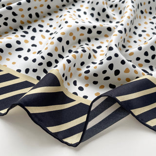 Silk scarf with white background featuring black and gold terrazzo dot pattern and navy striped border, elegantly draped to show texture