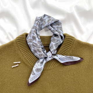 Pale blue silk bandana scarf with black paisley pattern tied elegantly around neck of mustard sweater, showcasing versatile styling option