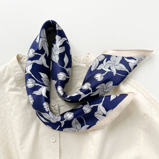 Navy blue silk bandana scarf with white floral pattern displayed elegantly around collar of cream blouse, showing shiny silk texture