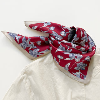 Vibrant red silk bandana scarf with blue and white botanical leaf pattern, casually draped around collar of cream garment, showcasing luxurious texture and elegant design