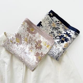 Two elegant Twilight Blossom silk scarves displayed on cream fabric - beige with mauve flowers and black-bordered with navy floral pattern, both featuring gold accents