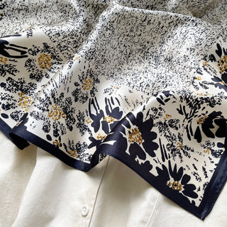 Elegant silk scarf with beige background featuring black floral patterns and gold accents, bordered with navy trim, draped over white fabric