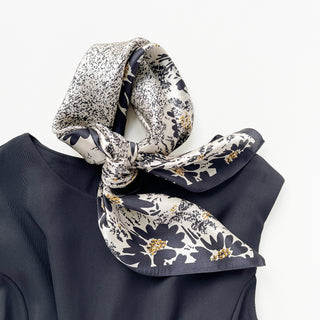 Elegant beige silk scarf with black floral pattern and gold accents, knotted stylishly on a navy blouse collar, showcasing luxurious texture and design.