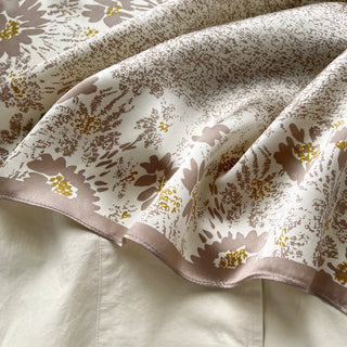 Beige silk scarf with delicate floral pattern featuring soft mauve blooms and gold accents, displaying luxurious fabric texture with gentle folds