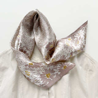 Beige silk scarf with mauve floral pattern and gold accents, elegantly tied on light-colored fabric, showcasing its luxurious sheen and delicate design.