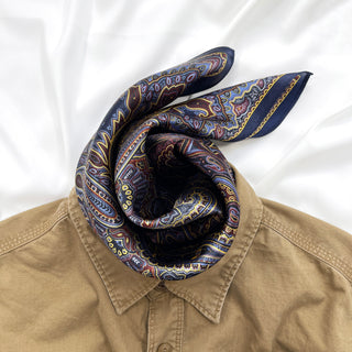 Navy blue silk neckerchief with intricate paisley pattern in burgundy and gold, styled on a tan shirt collar against white background