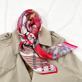 Magenta City Maze silk scarf with geometric patterns in red, pink, and blue, elegantly draped around the collar of a beige trench coat.