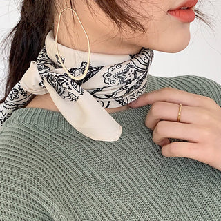 Light beige silk bandana scarf with black paisley pattern worn as a neck accessory with sage green sweater and gold hoop earring