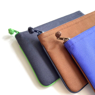 Three recycled RPET zipper wallets in gray with green trim, brown, and royal blue, featuring handmade button details and clean piped edges