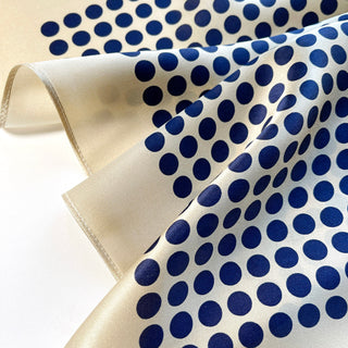 Luxurious silk bandana scarf with navy blue polka dots on light beige background, displaying smooth fabric texture and delicate hem edge